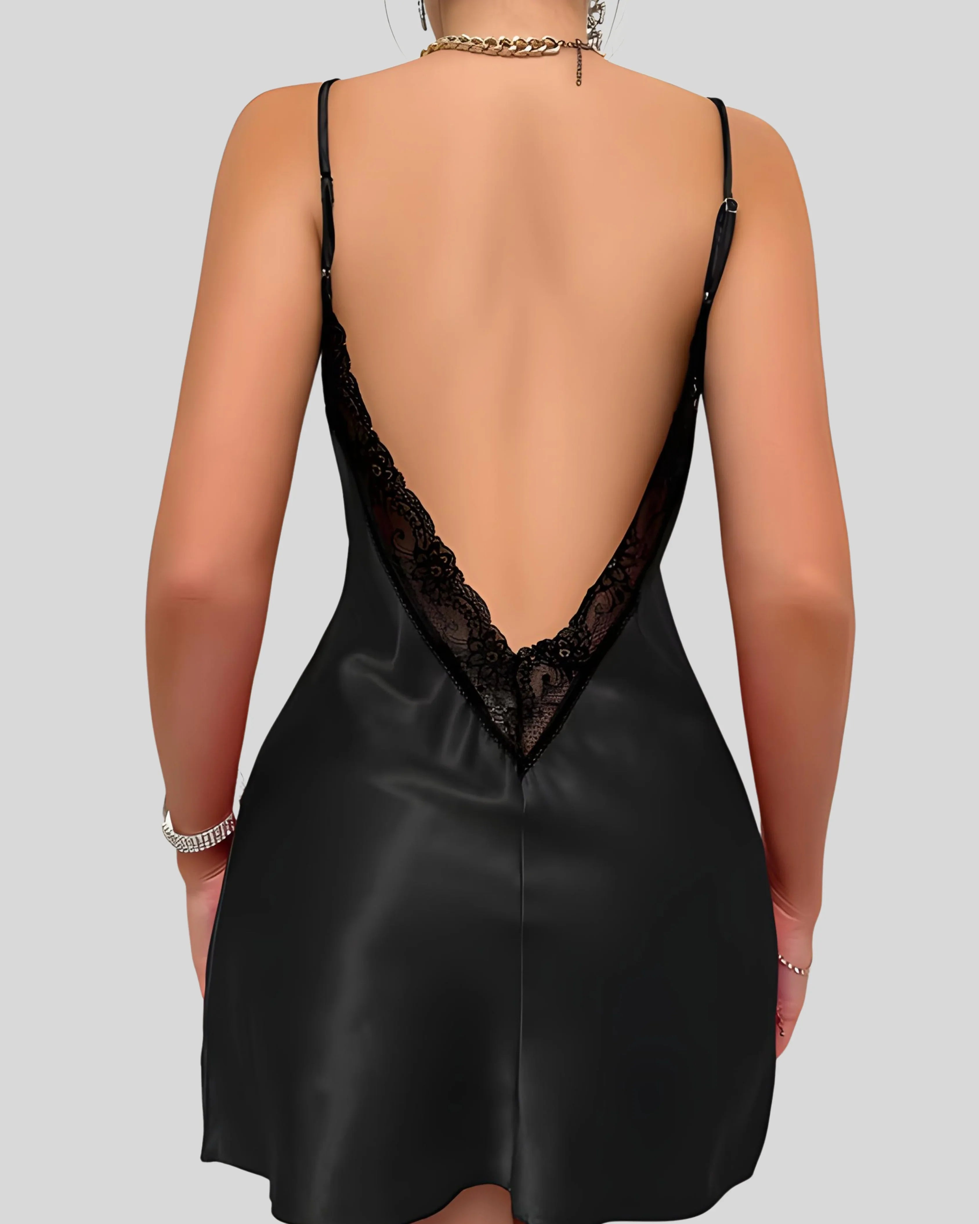 Woman wearing a black silk slip dress with thin straps, accessorized with gold chain necklace and bracelets on gray background